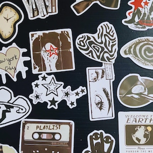 May include: A collection of various stickers with diverse designs. Includes a melting clock, a heart-shaped sticker with "new york city" text, a cassette tape labeled "PLAYLIST", and a sticker with the text "WELCOME TO EARTH". The stickers are in shades of brown, white, and black.