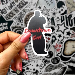 May include: A black and white sticker featuring a silhouette of a person with the words "Downtown Girl" in red. The sticker is held against a background of various black and white stickers with text and star designs.