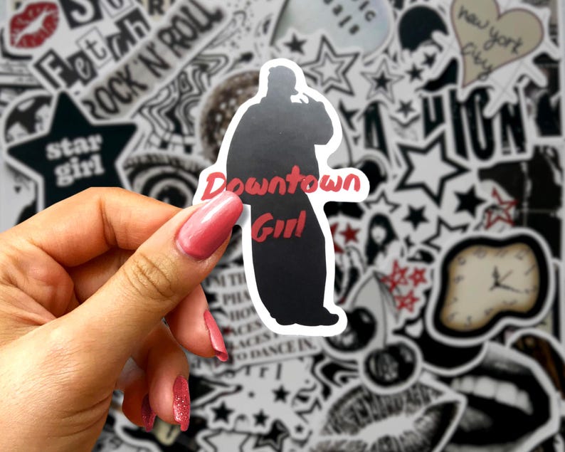 May include: A black and white sticker featuring a silhouette of a person with the words "Downtown Girl" in red. The sticker is held against a background of various black and white stickers with text and star designs.
