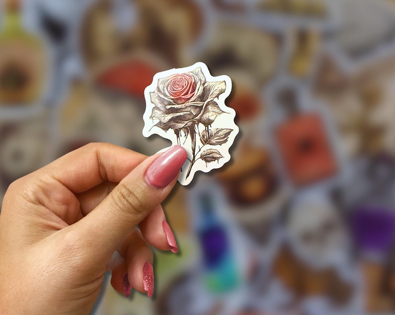 May include: A sticker featuring a detailed illustration of a rose with a red center and gray petals. The sticker has a white border and is held by a hand with pink nail polish. The background is blurred with other stickers.