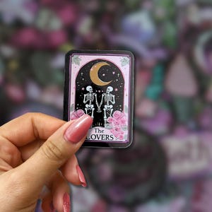 May include: A rectangular sticker featuring a tarot card design. The card depicts two skeletons holding hands under a crescent moon and stars, with pink roses at the bottom. The text "The Lovers" is at the bottom.