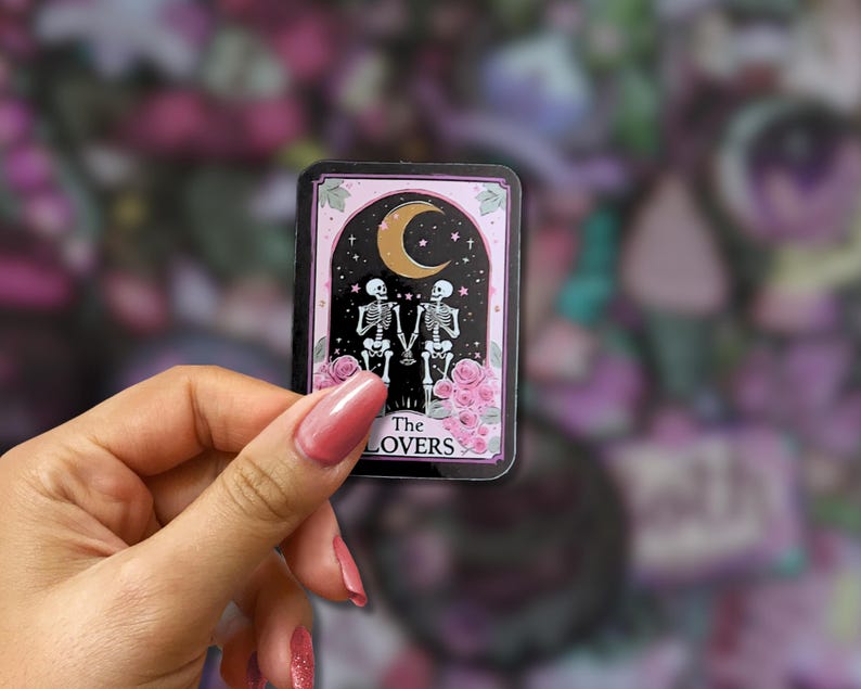 May include: A rectangular sticker featuring a tarot card design. The card depicts two skeletons holding hands under a crescent moon and stars, with pink roses at the bottom. The text "The Lovers" is at the bottom.