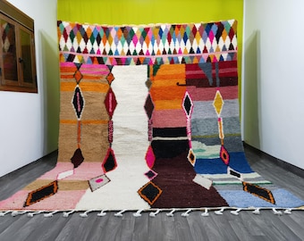 May include: A colorful Moroccan rug with a geometric pattern in shades of pink, orange, blue, and green. The rug has a white center with a diamond pattern and a fringe border.