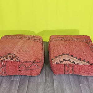 Può includere: Due cuscini da pavimento quadrati realizzati in tessuto rosso intrecciato con motivi geometrici marrone scuro. Ogni cuscino presenta una superficie superiore piatta e lati verticali dall'aspetto annodato e testurizzato, con motivi tribali.
