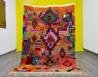 May include: A vibrant, hand-woven rug with a colorful geometric design. The rug features a mix of orange, pink, green, and blue hues, with a white fringe along the bottom edge. The intricate pattern includes diamond and square shapes.