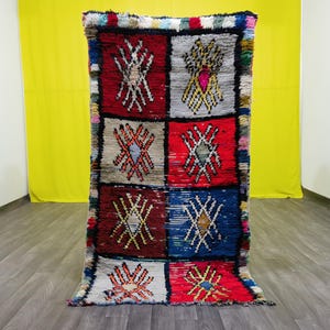 May include: A colourful Moroccan rug with a geometric pattern in shades of red, blue, white, and yellow. The rug is made of wool and has a hand-woven texture.