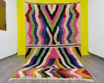 May include: A colorful Moroccan rug with a geometric pattern in shades of pink, green, blue, purple, and black. The rug has a white background and a fringe border.