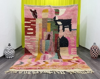 May include: A colorful, abstract Moroccan rug with a pink background and geometric shapes in red, yellow, green, and black. The rug has a fringed edge and is displayed on a wooden floor.