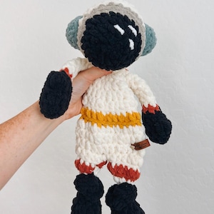 May include: Crocheted astronaut doll in a white spacesuit with black boots, gloves, and helmet. The helmet has a black faceplate with white accents. The suit features orange and red stripes. A small brown tag is attached to the doll.