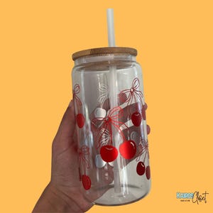 May include: A clear glass tumbler with a bamboo lid and a white straw. The tumbler is decorated with red cherries and white bows. The tumbler is held against a yellow background.