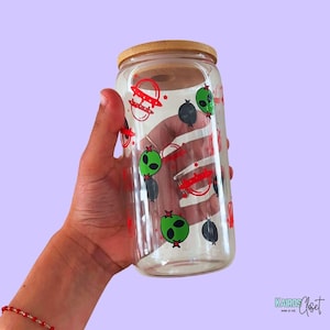 May include: A clear glass tumbler with a bamboo lid, decorated with green alien heads, red UFOs, and black planets. The tumbler is cylindrical and held in a hand against a light purple background. The design is repeated around the glass.