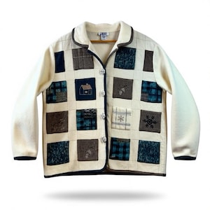 May include: Cream-colored fleece jacket with a patchwork design. The jacket features a collar, long sleeves, and a button-up front. The patchwork includes squares with embroidered images of a house, pinecones, and snowflakes. The jacket has a dark blue trim.