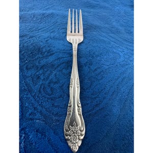 May include: A silver-toned metal dinner fork featuring a four-tined head and a handle decorated with embossed floral patterns and curved lines. The handle tapers toward the base, which is rounded and adorned with a detailed cluster of flowers.