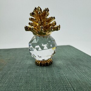 May include: A decorative pineapple figurine. The top is gold-colored, resembling a pineapple crown, and sits atop a faceted, clear crystal body. The base is also gold-colored. The figurine is displayed on a textured, green surface.