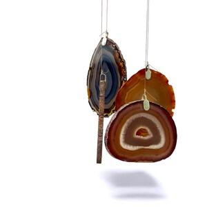 May include: A collection of agate slices in shades of brown, orange, and blue, suspended by thin wires. The agate slices have a polished appearance, showcasing natural patterns and textures. A wooden stick hangs from one of the slices.