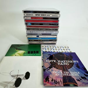 May include: A stack of music CDs featuring various artists and albums, including Spice Girls, Santana, and Dave Matthews Band. The CDs are in clear plastic cases, with album art visible. Some CD cases are stacked on top of each other.