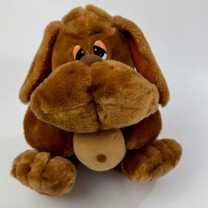 May include: A brown plush dog toy with floppy ears, a large snout, and a light brown tongue. The dog has large, expressive eyes with orange accents. The toy is sitting on a white surface.