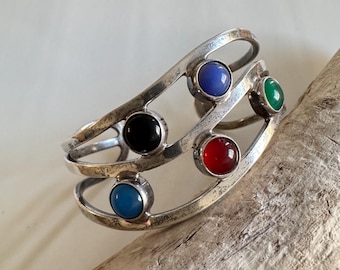 May include: A silver-toned cuff bracelet featuring six colorful, round gemstones. The gemstones are blue, black, red, and green. The bracelet has an open design with a curved shape.
