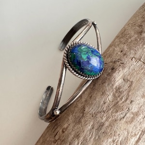 May include: A silver cuff bracelet with an oval stone in shades of blue and green. The stone is framed by a twisted rope-like border. The bracelet has an open design.