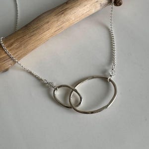 May include: A silver necklace with two interlinked oval pendants. The pendants are joined by a fine chain, with a small crystal near each pendant. The necklace is shown on a light surface.