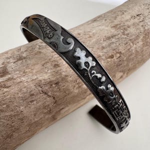 Puede incluir: Una pulsera de puño plateada con intrincados patrones florales y geométricos. La pulsera abierta presenta diseños en relieve detallados a lo largo de su superficie, incluyendo flores estilizadas y un fondo texturizado. El metal tiene una apariencia ligeramente envejecida.