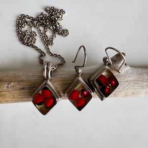 May include: A silver necklace and earring set. The pendant and earrings feature a diamond-shaped frame with red beads. The jewelry is displayed on a piece of driftwood against a white background.