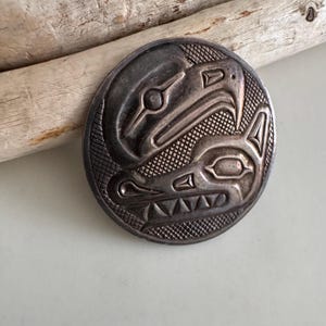 Peut inclure: Un pendentif rond en métal argenté avec un motif amérindien détaillé. Le pendentif présente deux figures d'animaux stylisées, peut-être un oiseau et un loup, sur un fond texturé. Le pendentif est présenté sur une surface de couleur claire.