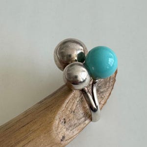 Pode incluir: Um anel de prata com três elementos esféricos: dois prateados e um turquesa. O anel é apresentado sobre um pedaço de madeira, destacando o contraste entre o metal e a textura natural.