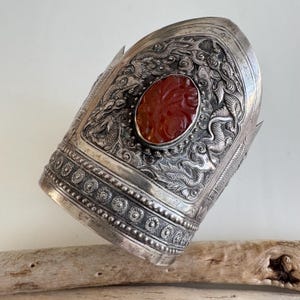 Puede incluir: Brazalete de puño plateado adornado con una gran piedra roja ovalada tallada. El brazalete está muy detallado con motivos de dragones y nubes, y patrones florales. El puño tiene una forma ancha y curvada.