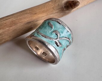 Puede incluir: Un anillo ancho de plata con un diseño de esmalte turquesa. El anillo presenta un patrón de desplazamiento en un tono más claro de turquesa, engastado sobre una banda de plata. El anillo mide aproximadamente 2,5 cm de ancho.
