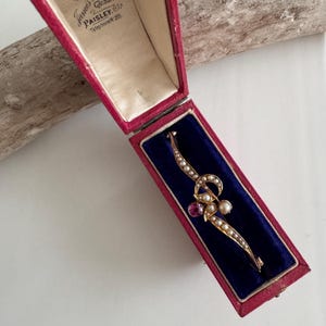 Puede incluir: Broche dorado vintage con un diseño curvo, adornado con pequeñas perlas y una gema rosa central. El broche se exhibe dentro de una caja de terciopelo rojo con el texto "James Paisley" en la tapa.