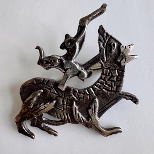 May include: A silver-toned brooch depicting a stylized wolf with an archer riding on its back. The wolf is in mid-stride, head raised, and the archer is aiming an arrow. The brooch has a detailed, textured design.