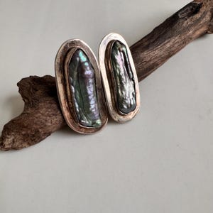May include: Two oval-shaped earrings with iridescent, dark gray-green pearls set in silver. The earrings are displayed on a piece of driftwood against a neutral background. The pearls have a unique, elongated shape.