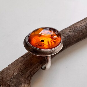 Könnte beinhalten: Silberring mit einem großen, ovalen Bernstein. Der Bernstein hat eine warme, orange Farbe mit sichtbaren Einschlüssen. Der Ring ist in einem schlichten, polierten Silberband gefasst.