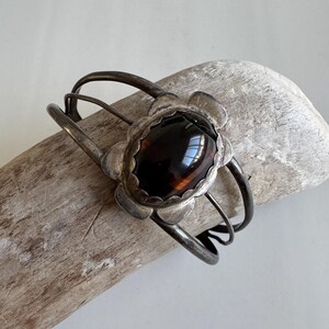 May include: A silver cuff bracelet featuring a large, oval, dark brown stone. The stone is set in a floral-shaped surround. The bracelet has a simple, open design with curved bands, suitable for a wrist.
