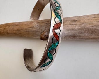 May include: A silver cuff bracelet with a turquoise and red inlay design. The bracelet has an open design and is resting on a piece of driftwood. The inlay pattern is a repeating series of linked shapes.
