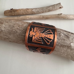 Pode incluir: Pulseira de punho de cobre com fundo preto e um design detalhado de Thunderbird. A pulseira está apoiada em um pedaço de madeira flutuante, mostrando seu intrincado trabalho em metal e estética de inspiração nativa americana.