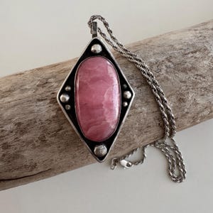 May include: A silver pendant necklace featuring a large, oval-shaped pink stone set within a diamond-shaped frame. The pendant hangs from a silver chain. The necklace is displayed on a piece of driftwood.