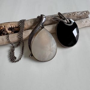 May include: Two silver necklaces with pendants. One pendant is a large, pear-shaped, off-white stone. The other is a black, oval-shaped stone. Both pendants are on silver chains. The necklaces are displayed on a piece of driftwood.