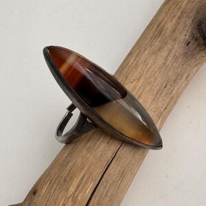 May include: A silver-toned ring with a large, elongated oval stone. The stone features bands of brown, amber, and cream colors. The ring is set on a piece of driftwood.