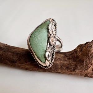 May include: A silver ring with a large, light green, triangular stone. The ring has floral and leaf accents and a twisted rope design. The ring is displayed on driftwood. The ring is likely made of silver.