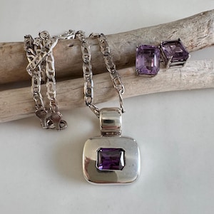 May include: A silver necklace with a square pendant featuring a purple gemstone, accompanied by matching square amethyst stud earrings. The jewelry is displayed on a piece of driftwood against a white background.