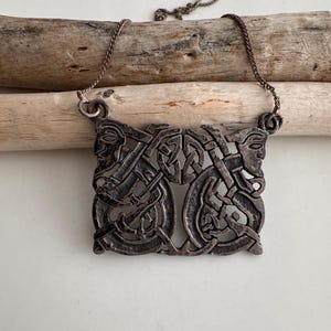 May include: A silver-toned pendant necklace featuring a rectangular pendant with an intricate Celtic knot design. The pendant showcases intertwined animal figures and geometric patterns. The chain is a delicate, matching silver color.