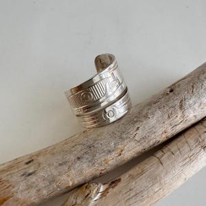 May include: A silver-colored ring with intricate engraved designs. The ring features a wrap-around style with detailed patterns, including lines and circular motifs. The ring is displayed on a piece of driftwood against a neutral background.