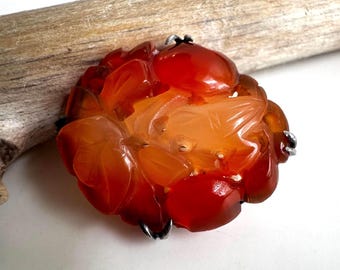 May include: A carved, orange-red flower brooch with a detailed floral design. The brooch has a silver-colored metal clasp and is set against a light background. The gemstone is translucent, with varying shades of orange and red.