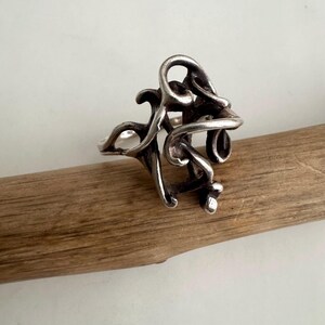May include: A silver-colored ring with an abstract design featuring swirling, organic shapes. The ring is set against a wooden background, showcasing its intricate details and unique form. The ring's design is reminiscent of flowing lines and curves.