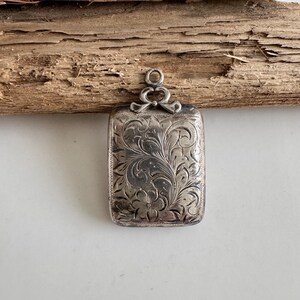 May include: A rectangular, silver-toned pendant with intricate floral and vine engravings. The pendant has a loop at the top with a decorative bow design. The surface is detailed with a vintage aesthetic.