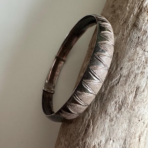 May include: A silver hinged bangle bracelet with a textured, chevron-like pattern. The bracelet has a clasp closure. The design features alternating smooth and textured sections, creating a visual contrast. The jewellery is displayed on a piece of driftwood.