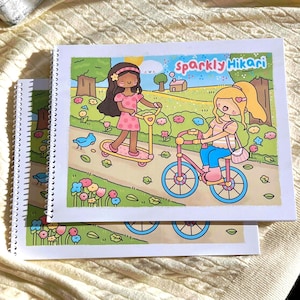 May include: Two spiral-bound notebooks with colorful illustrations. The top notebook features the text "Sparkly Hikari" and a cartoon scene of two girls, one on a scooter and the other on a bicycle, in a park setting.