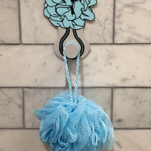 May include: A blue bath pouf hanging from a white and blue hook with a blue and white rope design. The hook is attached to a white tile wall.